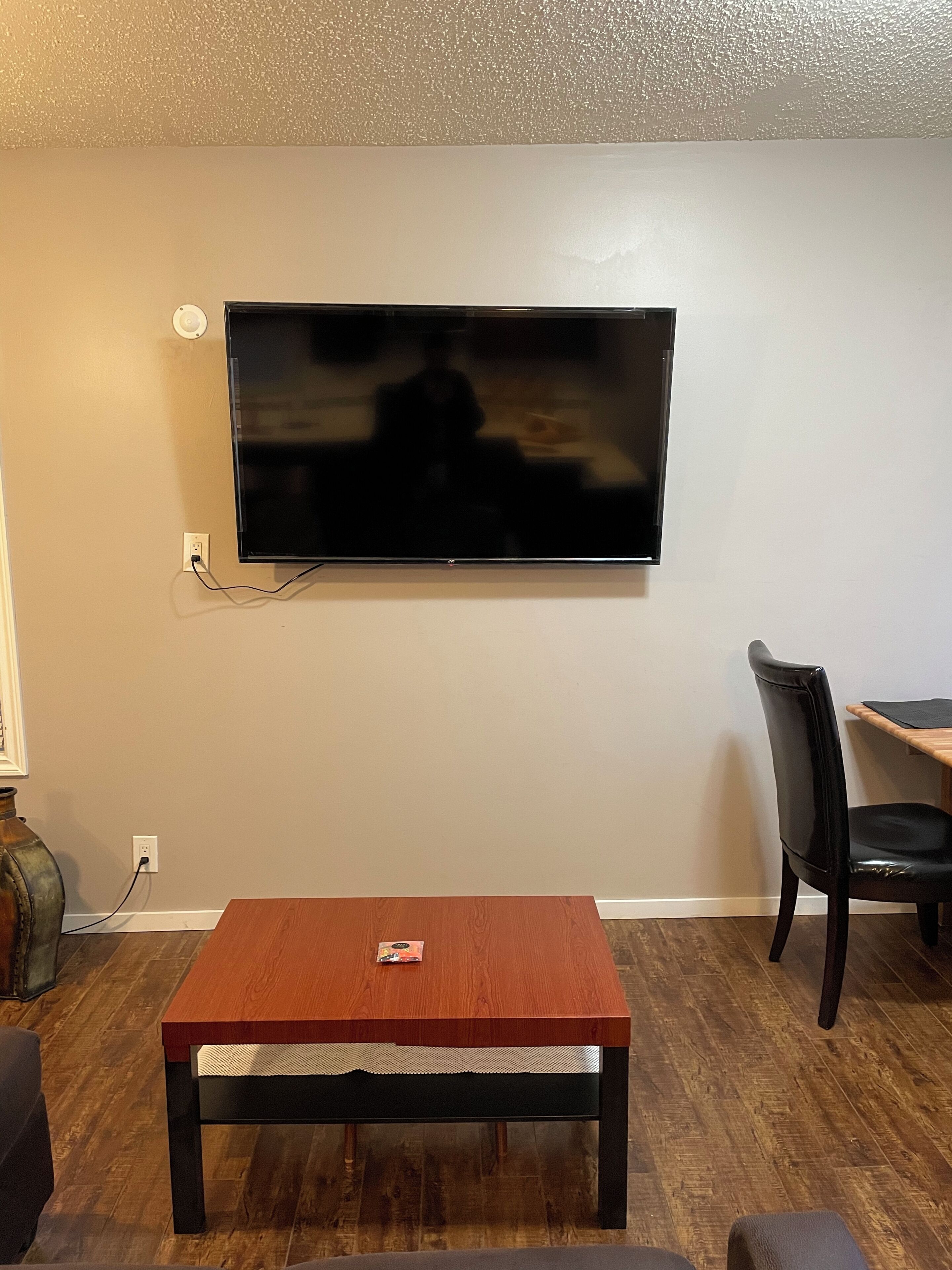 Een woonkamer met een flatscreen televisie aan de muur, een houten salontafel, een zwarte leren fauteuil en een klein tafeltje met een laptop.