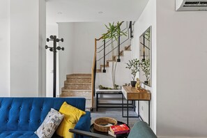 Un soggiorno moderno con un divano blu, un cuscino giallo e un tavolino da caffè in legno. Una scala con ringhiera in metallo conduce a una sala da pranzo con un tavolo e sedie in legno.
