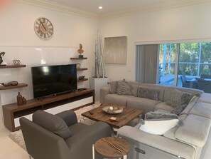 A modern living room with a flat-screen TV, a sofa, a coffee table, and a large window. - Island Time Starts Here! 3BR + Den Pool Retreat (Marco Island)