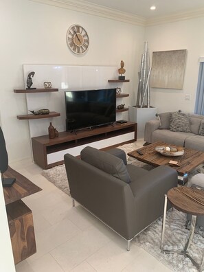 A modern living room with a flat-screen TV, a sofa, a coffee table, and decorative items on shelves. - Island Time Starts Here! 3BR + Den Pool Retreat (Marco Island)