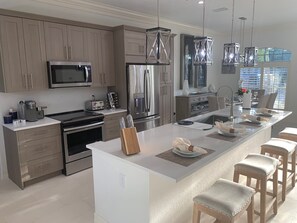 A modern kitchen with stainless steel appliances, a central island with bar stools, and pendant lighting. - Island Time Starts Here! 3BR + Den Pool Retreat (Marco Island)