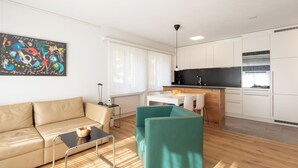 Uma sala de estar moderna com um sofá, uma mesa de centro e uma poltrona verde-água. Há uma área de cozinha com armários brancos e bancada preta.