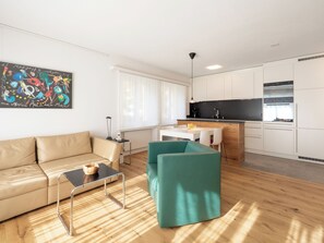 Ein modernes Wohnzimmer mit einer Couch, einem Couchtisch und einem türkisfarbenen Sessel. Es gibt eine Küchenzeile mit weißen Schränken und einer schwarzen Arbeitsplatte.