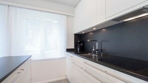 Uma cozinha moderna com bancada preta, armários brancos, forno embutido e janela com cortinas leves.