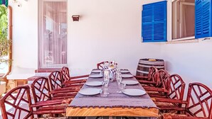 Uma sala de jantar com uma mesa comprida posta para várias pessoas, rodeada por cadeiras de vime, com uma janela e portaria azul na parede.