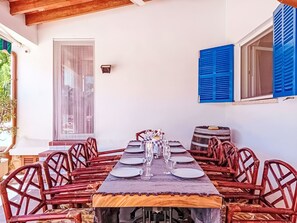 Un comedor con una larga mesa dispuesta para varias personas, rodeada de sillas de mimbre, con una ventana y contraventanas azules en la pared.