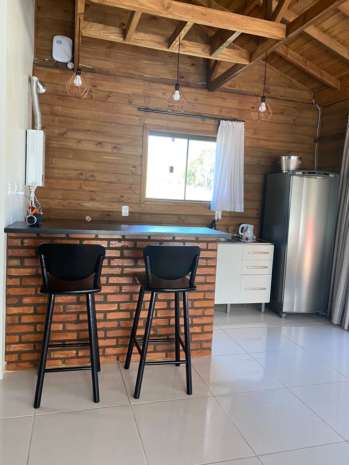 Una cocina con una isla de ladrillo, dos taburetes de bar, un refrigerador y lámparas colgantes.