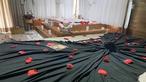 Une pièce avec un lit recouvert de draps noirs, de pétales de roses rouges et parsemé d’objets rouges en forme de cœur.