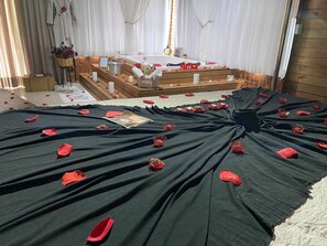 A room with a bed covered in black sheets, red rose petals, and scattered red heart-shaped objects.