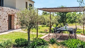 Um pátio com mesa e cadeiras, rodeado por um jardim com diversas plantas e um edifício ao fundo.