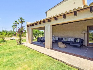 A covered outdoor seating area with a sofa and chairs, a lamp, and a view of a garden with palm trees and flowers. - Hort des Padri by Interhome (Marratxi)
