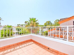 Ein Balkon mit weißer Brüstung, gefliestem Boden und Blick auf Palmen und Gebäude im Hintergrund.