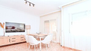 A modern dining area with a wooden table and white chairs, a television on the wall, and a lamp on a cabinet. - Olivos 3 by Interhome (Los Urrutias)