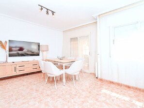 A modern dining area with a wooden table and white chairs, a television on the wall, and a lamp on a cabinet. - Olivos 3 by Interhome (Los Urrutias)