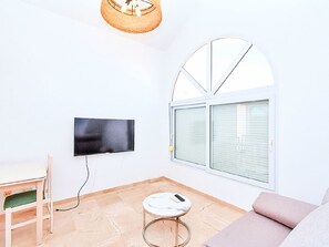 Ein Zimmer mit Flachbildfernseher an der Wand, einem runden Tisch mit Fernbedienung, einer Couch und einem Fenster mit Jalousien.
