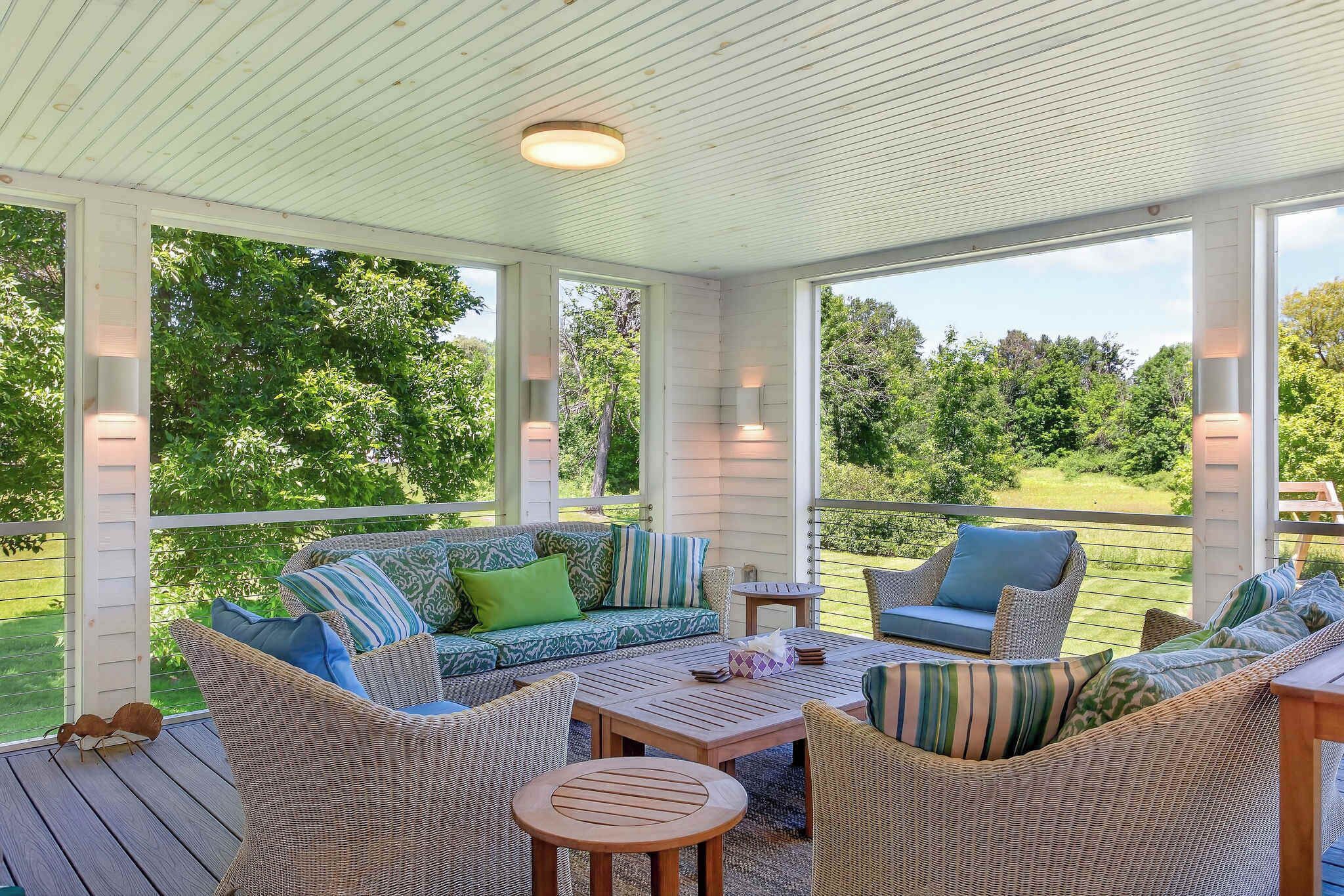 A covered outdoor seating area with wicker furniture, a wooden table, and a view of a green landscape.