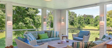 A covered outdoor seating area with wicker furniture, a wooden table, and a view of a green landscape.