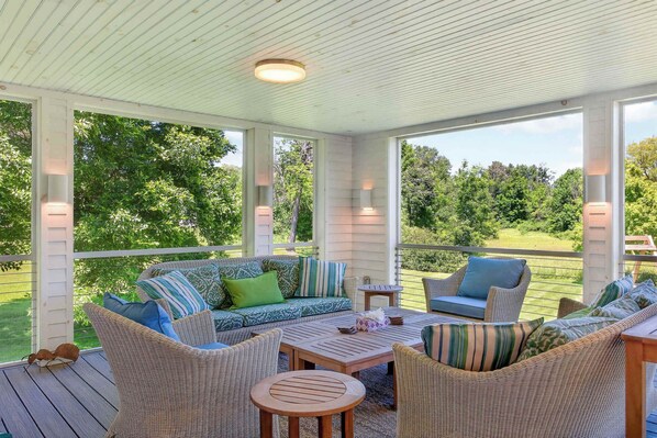 A covered outdoor seating area with wicker furniture, a wooden table, and a view of a green landscape.