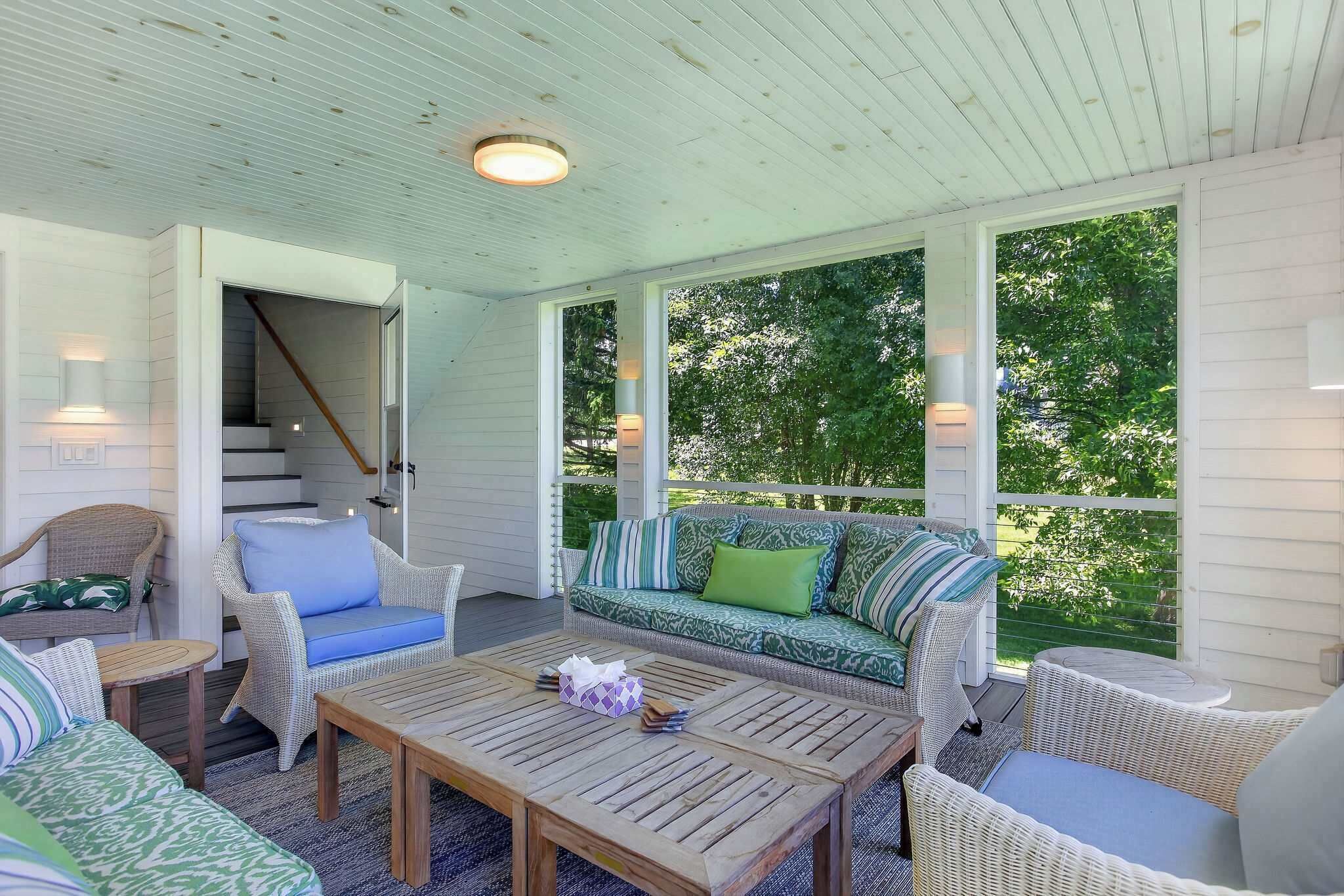 A covered outdoor seating area with wicker furniture, a wooden coffee table, and a staircase leading to an upper level.