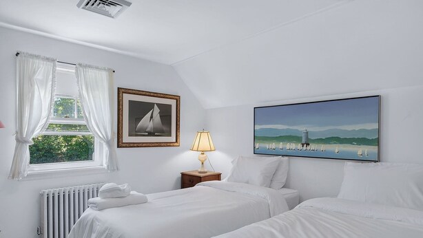 Quarto com duas camas, um radiador, janela com cortinas, mesa de cabeceira com abajur e duas fotos emolduradas na parede.