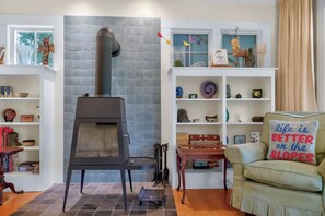 Una sala de estar acogedora con una chimenea de leña, estantes con objetos decorativos y un sillón con cojín.