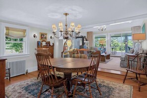 A dining area with a round table and chairs, a chandelier, and a view of the outdoors through large windows.