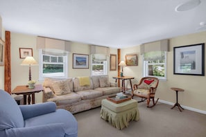 A living room with a sofa, armchair, ottoman, and a rocking chair. There are two windows with curtains, a lamp, and framed pictures on the walls.