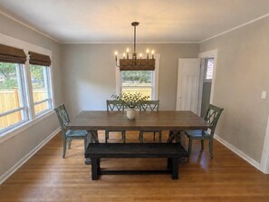 A dining room with a large wooden table, six chairs, a bench, a chandelier, and a window with a view of greenery. - Spacious Coastal-Style Home in Bryant — Steps to UW, U-Village Mall, and more! (Seattle)