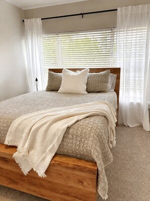 Quarto com cama de madeira, roupa de cama branca e cortinas leves.