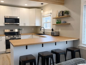 A modern kitchen with white cabinets, a stainless steel microwave, a built-in oven, a wooden countertop, a sink, and a toaster. There are three stools and a window with blinds. - Spacious Coastal-Style Home in Bryant — Steps to UW, U-Village Mall, and more! (Seattle)