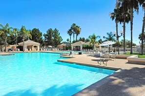 A swimming pool with lounge chairs and umbrellas, surrounded by palm trees and covered structures.