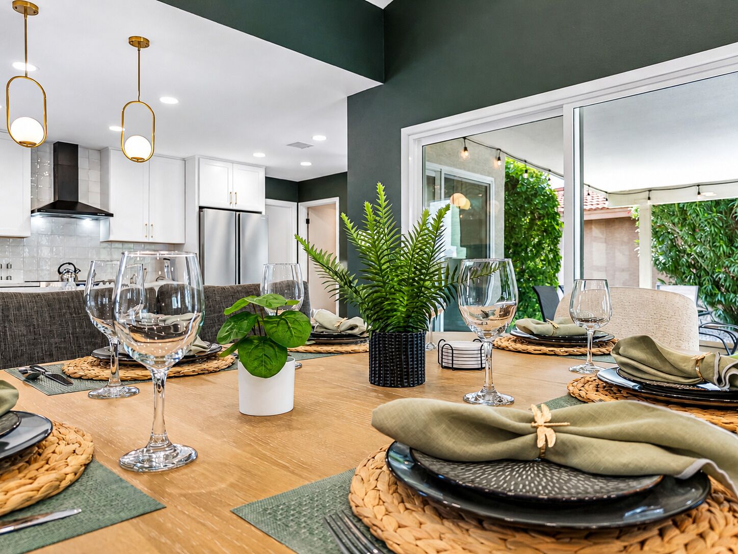 A modern dining area with a wooden table set for a meal, surrounded by kitchen cabinets and a large window with a view of greenery.