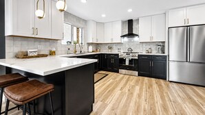 Una cucina moderna con mobili bianchi, un'isola centrale con sgabelli, elettrodomestici in acciaio inossidabile e illuminazione a sospensione.