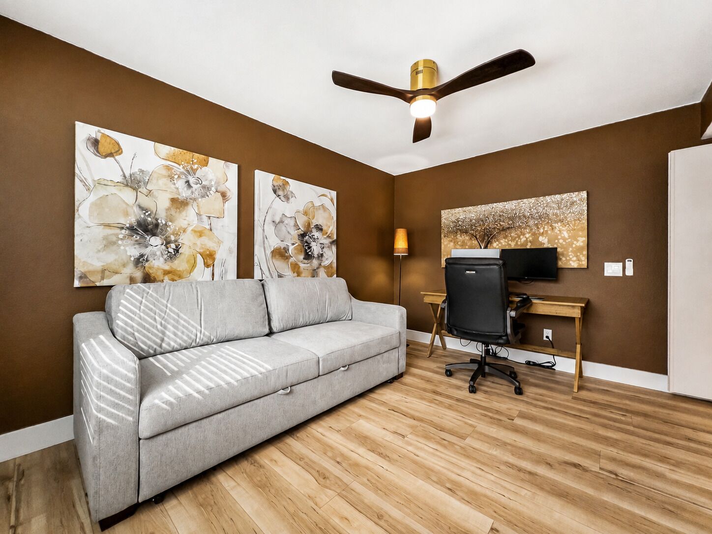 Un salon moderne avec un canapé gris, un plancher en bois, un ventilateur de plafond et des œuvres d’art accrochées aux murs.