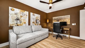 Un salon moderne avec un canapé gris, un plancher en bois, un ventilateur de plafond et des œuvres d’art accrochées aux murs.