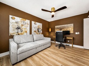 A modern living room with a grey sofa, wooden flooring, a ceiling fan, and artwork on the walls.