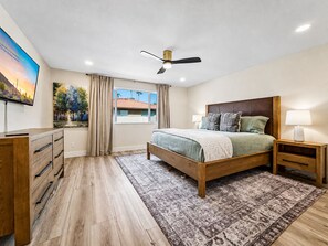 A bedroom with a bed, bedside tables, a ceiling fan, a window with curtains, and a painting on the wall.
