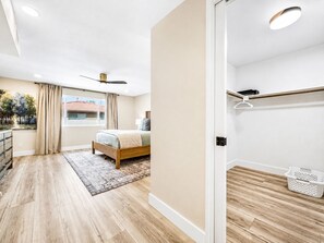 Un dormitorio con cama, ventilador de techo, ventana con cortinas, suelo de madera y vista a un edificio.
