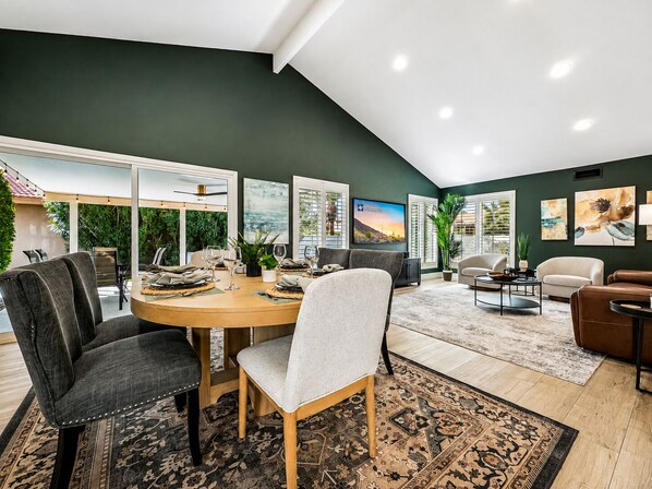 A modern living room with a dining area, large windows, and a view of greenery.