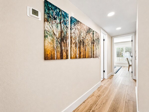 A hallway with wooden flooring, a thermostat, and two abstract paintings on the wall.