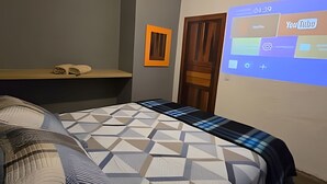 Un dormitorio con cama, mesita de noche, proyector montado en la pared y una puerta.