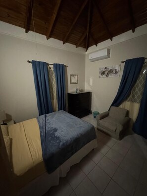 A bedroom with a bed, a chair, a small table, and an air conditioning unit. - Mansfield Elegance (Ocho Rios)