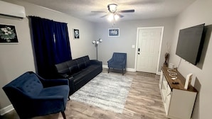 Un salon avec un canapé noir, un fauteuil bleu, un meuble en bois et un téléviseur fixé au mur.