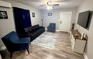 A living room with a black sofa, a blue armchair, a wooden cabinet, and a television on the wall. - 2-bedroom apartment in peaceful Houston with AC for relaxing stays (Houston)