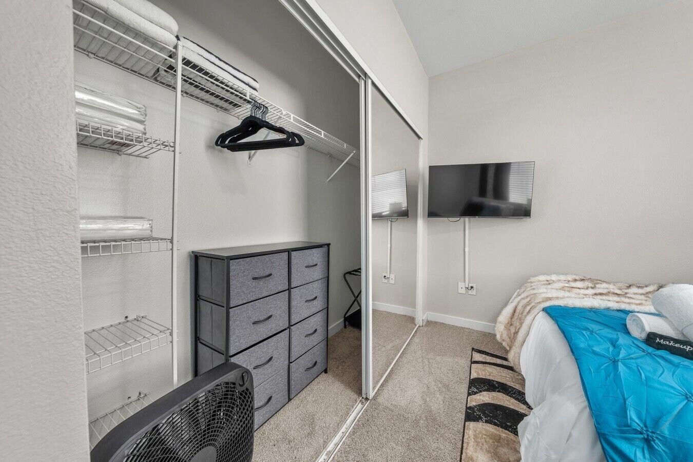 A bedroom with a bed, a dresser, a closet with shelves, a TV, and a fan.