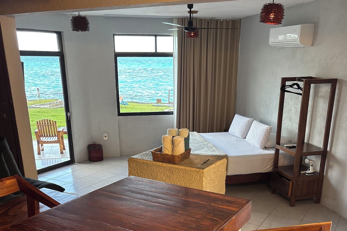 A hotel room with a wooden dining table, a bed with white linens, a hanging light fixture, and a view of the sea through a large window.