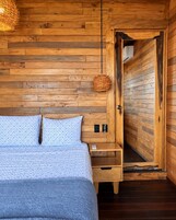 Quarto de cabana de madeira com cama, mesa de cabeceira e porta que dá para outro cômodo.