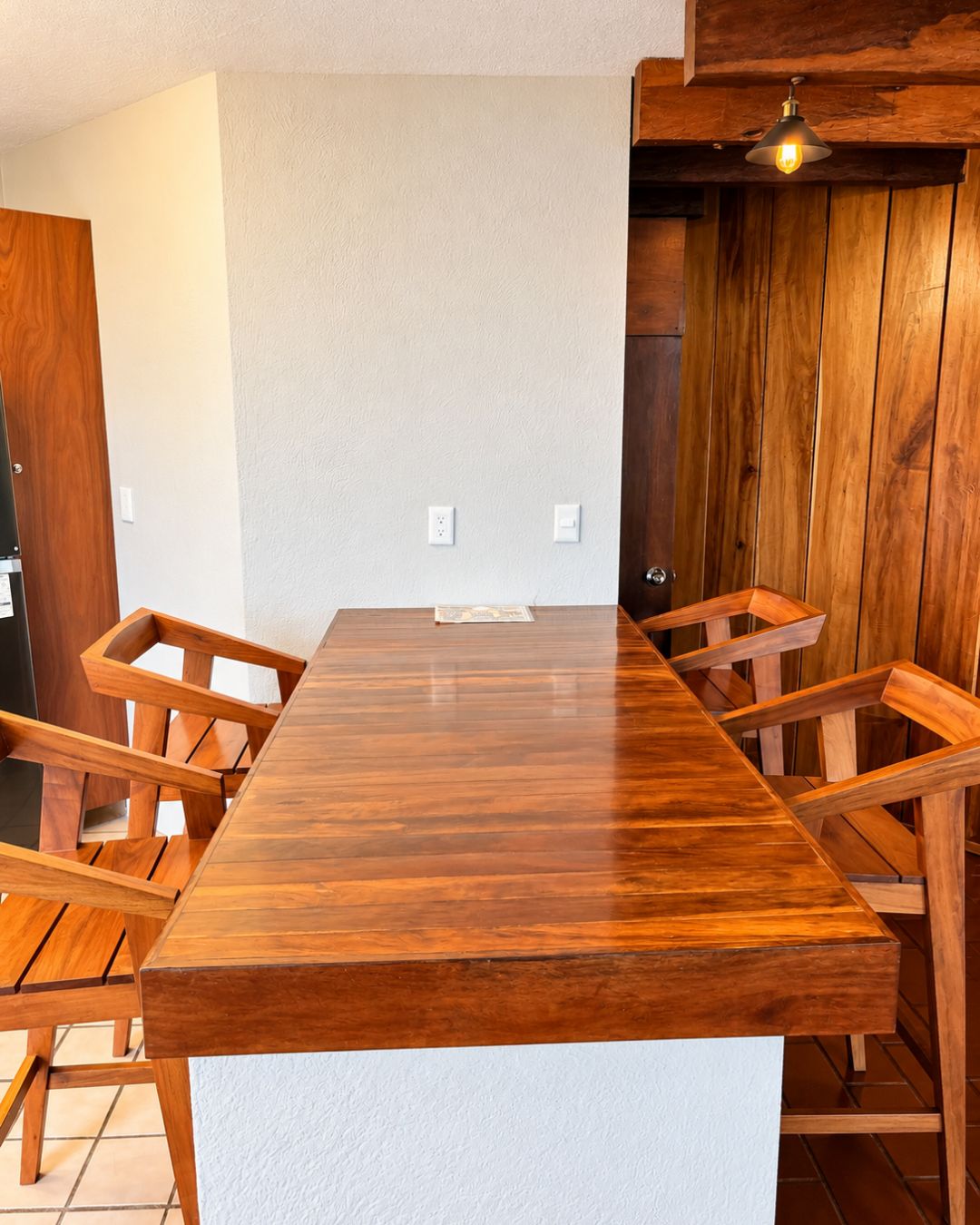 A wooden dining table with six chairs, a white wall, and a wooden ceiling with a light fixture.