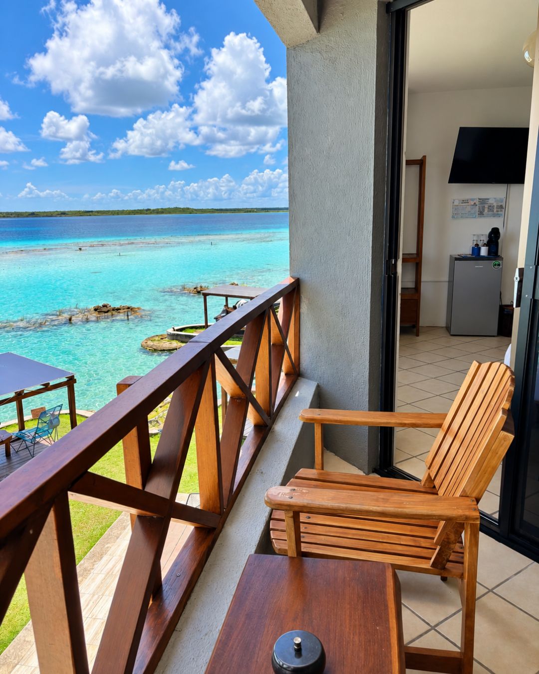 A balcony with a wooden chair and table, overlooking a turquoise sea and clear sky.