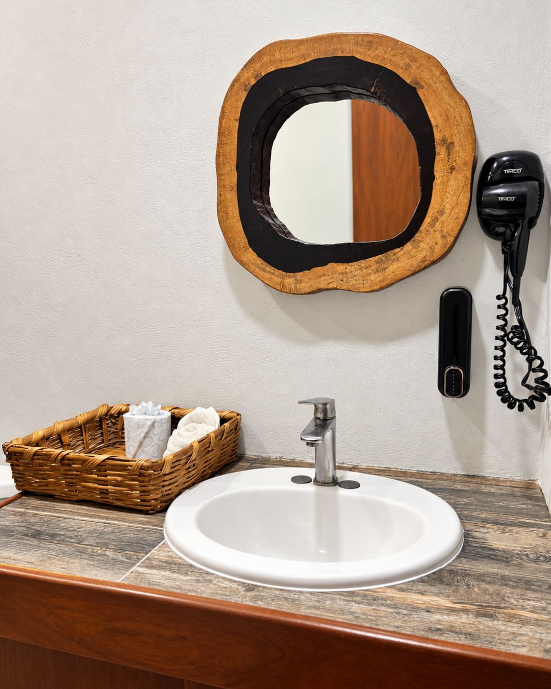 Un baño con un espejo de marco de madera, un lavabo blanco, un teléfono negro y una cesta de mimbre con toallas.